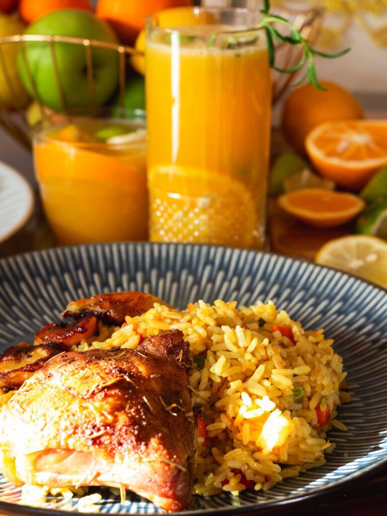 Plate of Nigerian coconut rice and chicken and fried plantain with juice in the background